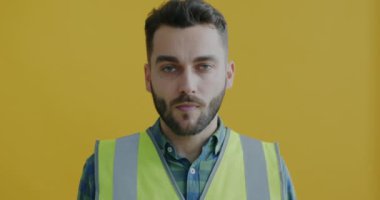 Yelekli profesyonel ressamın portresi. Sarı arka planda bir kavanoz duvar boyası ve silindir ile el kaldırıyor. Onarım ve enstrüman konsepti.