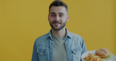 Sağlıklı sebze salatası yerine fast food burger ve patates kızartmasını seçen ve sarı arka planda gülümseyen genç adamın portresi. Gençlik yaşam tarzı ve beslenme konsepti.
