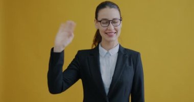 Resmi takım elbiseli, el sallayan ve sarı arka planda gülümseyen iş kadınının portresi. İfade ve olumlu duygu kavramı.