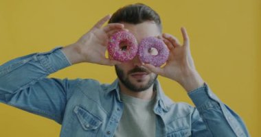 Donutlarla eğlenen, şeker bardakları yapan ve sarı arka planda suratını buruşturan oyuncu portresi. İnsanlar ve tatlı yemek konsepti.