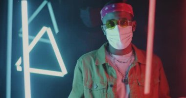 Neon ışıklı arka planda yüz maskesi takan mutsuz Afrikalı Amerikalı adam yavaş hareket ediyor. İnsanlar ve salgın konsepti.