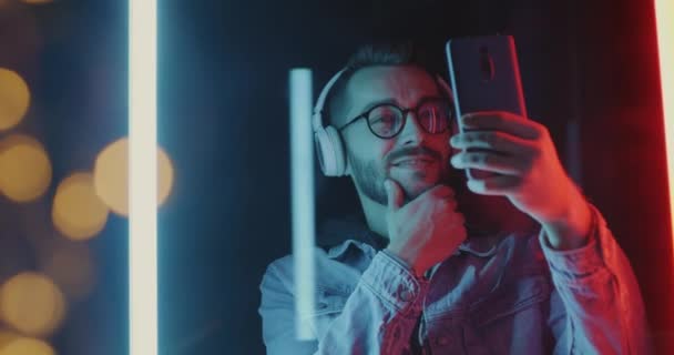 Portret Faceta Słuchawkami Robiącego Selfie Smartfonem Pokazującym Kciuki Góry Neonowym — Wideo stockowe