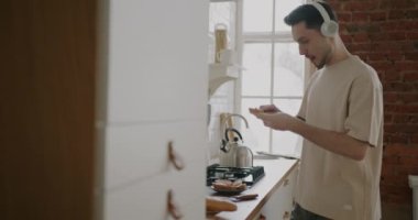 Evde mutfakta dans eden ve kulaklıkla müzik dinleyen genç bir adamın portresi. Gıda hazırlama ve insan konsepti.