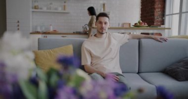 Karısı mutfakta yürürken, evdeki kanepede oturan sakin genç adamın yavaş çekimde portresi. Aile hayatı ve iç mekan kavramı.