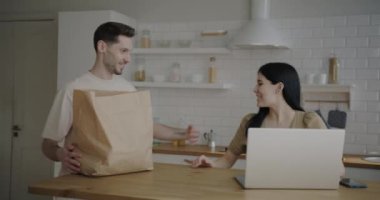 Kadın dizüstü bilgisayarla çalışırken, adam mutfağa erzak getiriyor ve karısıyla konuşurken paketleri açıyor. Ev hayatı ve alışveriş konsepti.