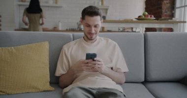 Kadın mutfakta yürürken akıllı telefon kullanan genç bir adam evdeki koltukta oturuyor. Aile hayatı ve eğlence etkinliği kavramı.