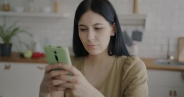 Ciddi bir genç kadın akıllı telefon mesajı kullanıyor ve tek başına mutfakta sergileniyor. Çevrimiçi iletişim ve modern teknoloji kavramı.