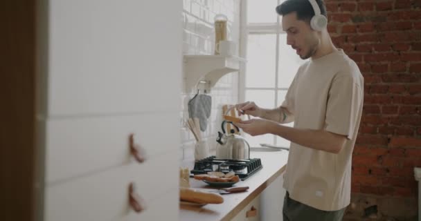 Веселый Человек Готовит Сэндвич Наслаждаясь Музыкой Через Наушники Танцуя Кухне — стоковое видео