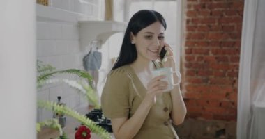 Tasasız bayan cep telefonuyla konuşuyor ve mutfaktaki kupadan çay içiyor. Modern teknoloji ve cep telefonu bağlantısı kavramı.
