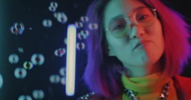 Oyuncu Asyalı kadın sabun köpüğü oyuncak tabancasıyla eğleniyor ve parlak neon arka planda dans ediyor. Neşeli yetişkin eğlencesi ve eğlence etkinliği konsepti.