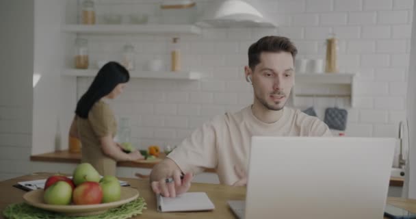 年轻男子在家里用笔记本电脑进行在线视频通话 而妻子则在家里做背景午餐 家庭生活和商人概念 — 图库视频影像