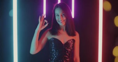 Tatmin olmuş genç bir bayanın portresi. Tamam el hareketi yapıyor ve gülümsüyor. Güzel siyah arka planda, neon lambaları ve bokeh ışıklarıyla duruyor.