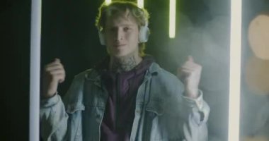 Kulaklık takan, siyah arka planda yanıp sönen neon ışıklarla dans eden şık bir gencin yavaş çekim portresi. Gençlik ve aletler konsepti.