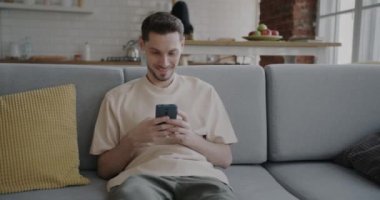 Kaygısız koca, evde yemek pişirirken akıllı telefondan gülümseyerek sosyal medyada eğleniyor. İnsanlar ve modern teknoloji kavramı.