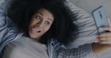 Afro-Amerikalı bir kızın evde yatakta yatarken akıllı telefonuyla selfie çekmesi. Modern teknoloji ve eğlence kavramı.