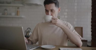Dizüstü bilgisayarla çalışan ve mutfakta kahve içen genç iş adamının portresi. Uzak çalışma ve gençlik yaşam tarzı konsepti.