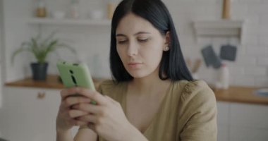Akıllı telefon mesajları kullanan düşünceli genç bir bayanın portresi mutfaktaki görüntülere bakıyor. Çevrimiçi iletişim ve aygıt kavramı.