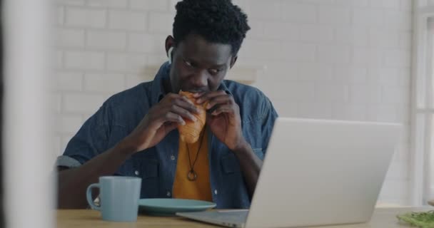 楽しいアフリカ系アメリカ人の男性は 自宅のキッチンでラップトップ画面を見てクロワッサンを食べて笑っています 現代のライフスタイルと国内レジャー活動のコンセプト — ストック動画