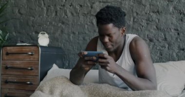 Evdeki yatakta akıllı telefonuyla online video oyunu oynayan neşeli Afro-Amerikalı adamın portresi. Modern teknoloji ve e-spor kavramı.