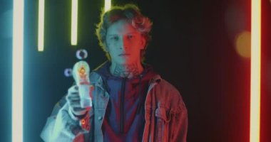 Ciddi bir genç adamın portresi oyuncak tabanca ateşliyor sabun köpükleri yapıyor koyu renkli neon arka planda dalgın yüzlü köpüğü seyrediyor. Eğlenceli yetişkin ve eğlence konsepti.