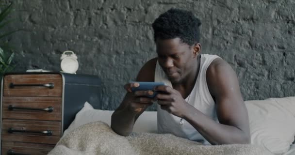 自宅でベッドでスマートフォンでオンラインビデオゲームをプレイ陽気なアフリカ系アメリカ人男性の肖像画 現代技術とEスポーツの概念 — ストック動画