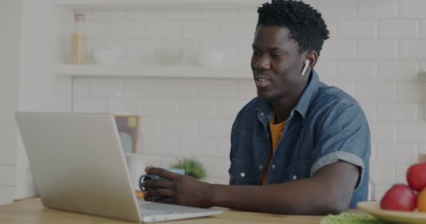 アフリカ系アメリカ人の男性がノートパソコンやイヤホンを使ってビデオ通話をしたり 自宅のキッチンで手を振っている姿 人とコミュニケーションの概念 — ストック動画