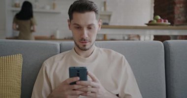 Neşeli adam akıllı telefon kullanır ve evde gülümserken karısı arka planda mutfakla meşgul olur. Aile yaşam tarzı ve modern teknoloji kavramı.