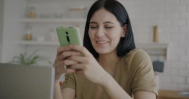 Akıllı telefondan gülümseyen neşeli genç bir bayanın portresi mutfaktaki sosyal medyadan keyif alıyor. Modern teknoloji ve iletişim konsepti.