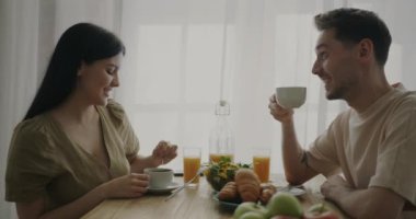 Genç ve mutlu bir çift kahvaltı yapıyor, kahve içiyor ve sohbet ediyorlar. Evdeki mutfakta masada oturuyorlar. İlişki ve yemek kavramı.