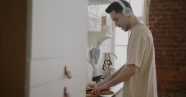 Kulaklıkla müzik dinleyen ve mutfakta sandviç yapan genç bir adamın portresi. Gıda hazırlama ve rahatlama konsepti.