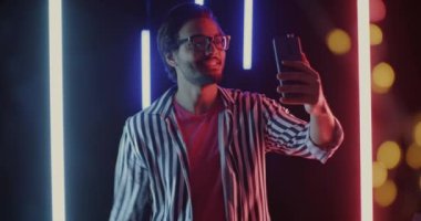 Çevrimiçi video görüşmesi yapan neşeli Orta Doğulu birinin portresi akıllı telefona işaret ederek neon arka planda baş parmağını gösteriyor. İletişim ve gece hayatı kavramı.