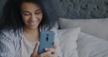 Akıllı telefon kullanıp ekranlara dokunan ve yatakta gülümseyen neşeli kadının portresi. Çevrimiçi iletişim ve modern teknoloji kavramı.