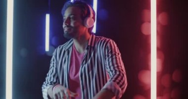Kulaklıklı mutlu Orta Doğulu öğrencinin yavaş çekimde güzel arka planda neon lambaları ve bokeh ışıkları ile dans edişinin portresi. Genç adam dinleniyor ve tek başına müzik dinliyor..