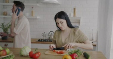 Genç bir kadın salata hazırlarken kocası cep telefonuyla konuşuyor ve evde sebze yiyor. Halk ve aile hayatı kavramı.