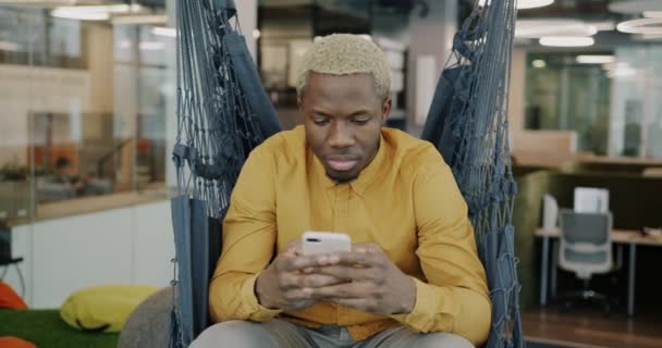 非裔美国人正在使用智能手机触摸屏放松在舒适的吊床在创造性的办公室 现代装置和工作概念 — 图库视频影像