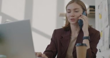 Kendine güvenen iş kadınının portresi cep telefonuyla konuşuyor ve iş yerinde dizüstü bilgisayar kullanıyor. Mobil teknoloji ve iletişim konsepti.