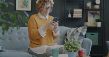 Modern akıllı telefon kullanan ve dairede fincandan çay içen neşeli genç bir kadının portresi. Boş zaman ve içecek konsepti.