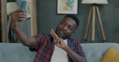 Akıllı telefon kamerasına poz veren neşeli genç adam evde el hareketi yaparken selfie çekiyor. Teknoloji ve sosyal medya kavramı.