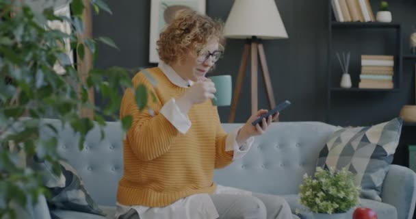 Mutlu Öğrenci Fincandan Kahve Içiyor Oturma Odasında Akıllı Telefonla Eğleniyor — Stok video