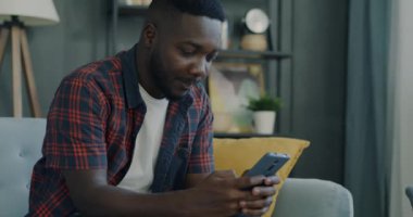 Gülümseyen Afro-Amerikan adam akıllı telefon kullanıyor evde mesajlaşıyor. Elektronik cihaz ve online iletişim konsepti.