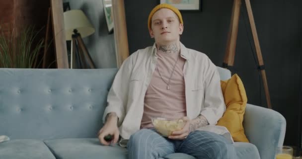 一个快乐的年轻人 带着纹身看电视 坐在沙发上吃爆米花 独自享受闲暇时光 娱乐和青年生活方式概念 — 图库视频影像