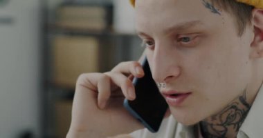 Piercingli ve dövmeli genç bir adamın yakın plan portresi. Ofisten cep telefonuyla iş görüşmesi yapıyor. İletişim ve insan kavramı.