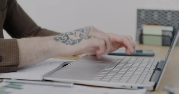 机の上でノートパソコンで作業を入力して入れ墨を持つ男性の手のクローズアップ 仕事に集中した現代のコンピュータを使用して創造的な男 — ストック動画