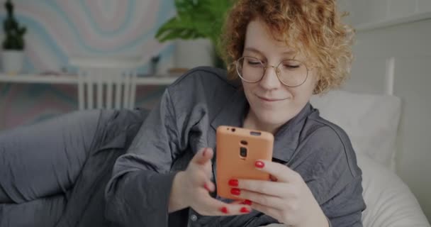 Evde Yatakta Gülümseyen Akıllı Telefon Kullanan Pijamalı Neşeli Kızıl Saçlı — Stok video