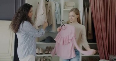 Moda butiğindeki müşterilere kıyafet seçmede yardımcı olan tezgahtar. Sarışın kadın bluz alıyor, elinde moda kıyafetler, gülümsüyor..