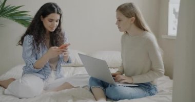 Diz üstü bilgisayar, akıllı telefon ve yatak odasında oturan mutlu genç bayan arkadaşlar. Modern teknoloji ve arkadaşlık kavramı.
