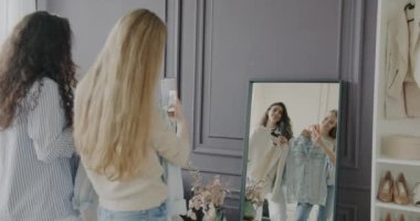 Neşeli kızlar kıyafet seçer ve modern butikte aynada akıllı telefonla fotoğraf çekerler. Alışveriş ve arkadaşlık kavramı.