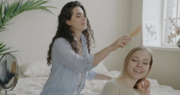 若い女性は姉妹の髪をブラッシングし 自宅で寝室で一緒に余暇時間を楽しんで話しています 美と家族関係の概念 — ストック動画