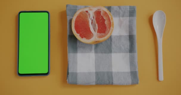 緑の画面のクロマキースマートフォンと女性の手のフラットレイアウト黄色の背景にグレープフルーツの半分を置く 現代の技術と果物の概念 — ストック動画