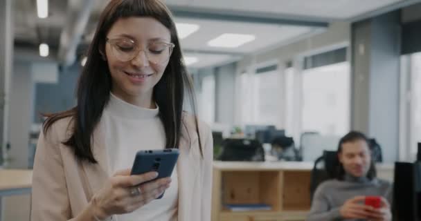 オフィスを歩いている楽しい女性社員のドリーショットやスマートフォンでテキストメッセージを送る一方で 同僚の多様なグループがバックグラウンドで働いています — ストック動画